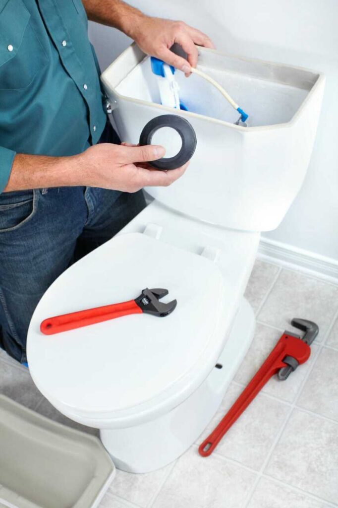 Person repairing a toilet tank with plumbing tools on the toilet seat and floor.