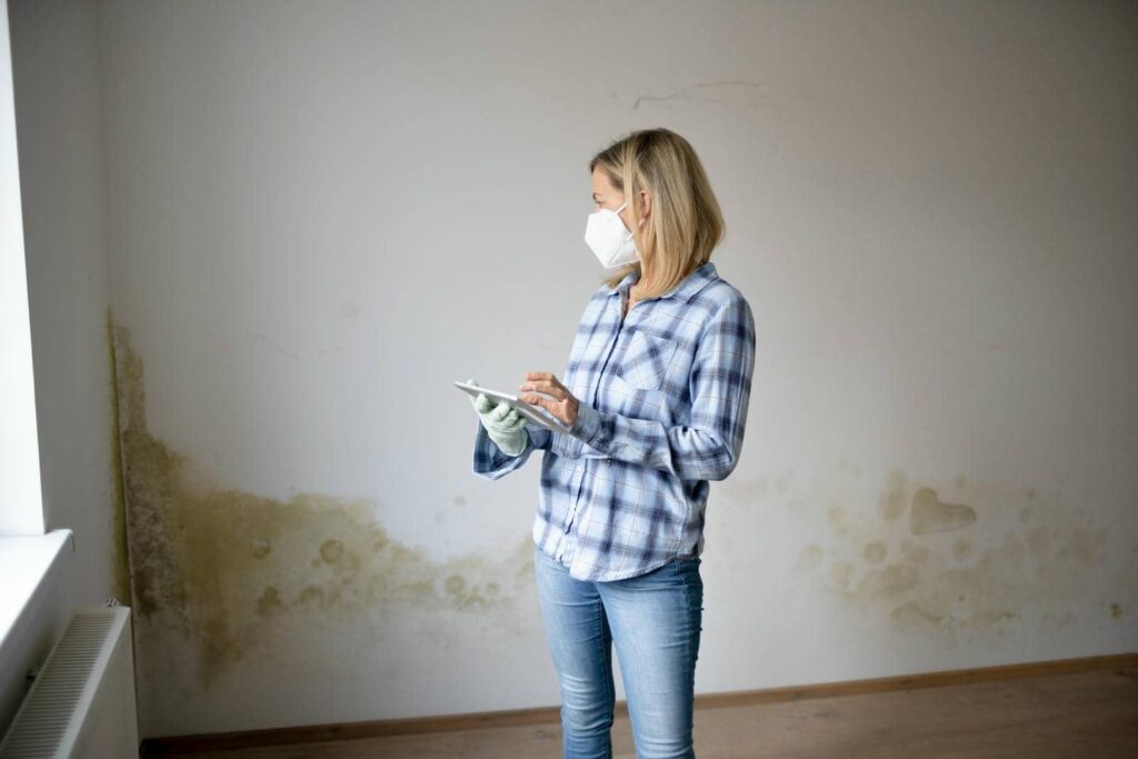 Woman wearing a mask and gloves inspecting mold damage on an interior wall while using a tablet.