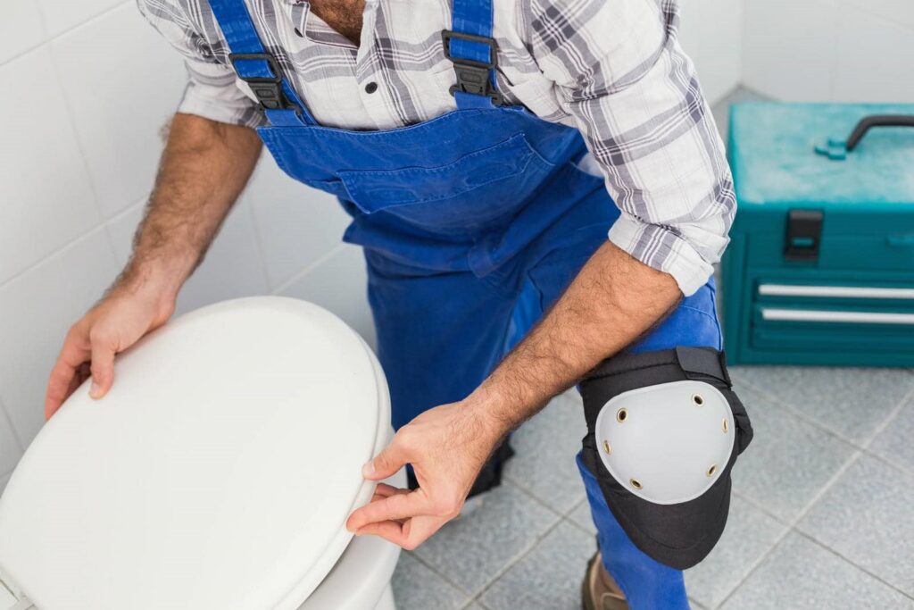 Plumber in blue overalls fixing a toilet seat in a tiled bathroom.