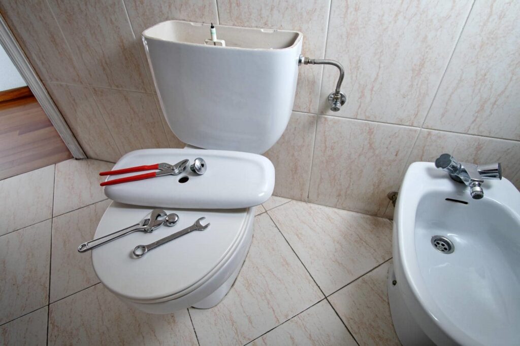 Toilet with open tank and tools on the seat and lid in a tiled bathroom.
