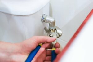 Hand using a wrench to tighten a metal pipe fitting near a toilet.